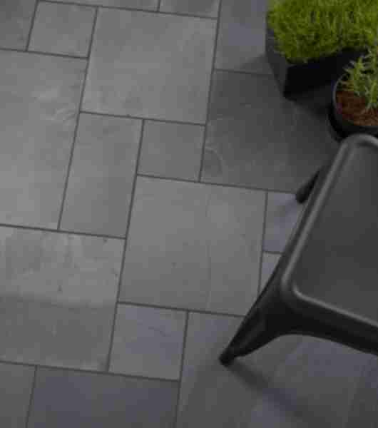 Outdoor patio paved with black slate Versailles patterned tile. A chair and plants are arranged on the patio.