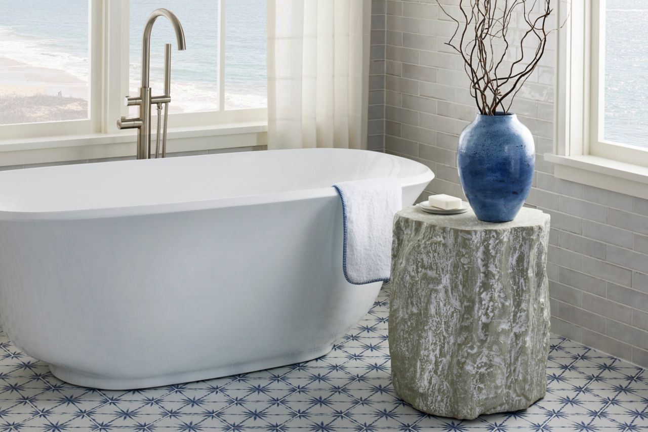 White tub in a bathroom with large windows and blue and white porcelain hexagon tile on the floor. Small table with decorative branches in a blue vase and soap.