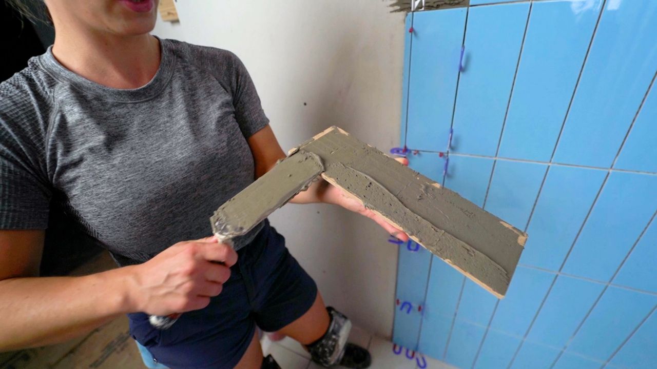 A person using a trowel to apply thinset mortar to the back of a piece of tile, focusing on applying extra mortar in the places where a metal profile piece will sit underneath the tile.