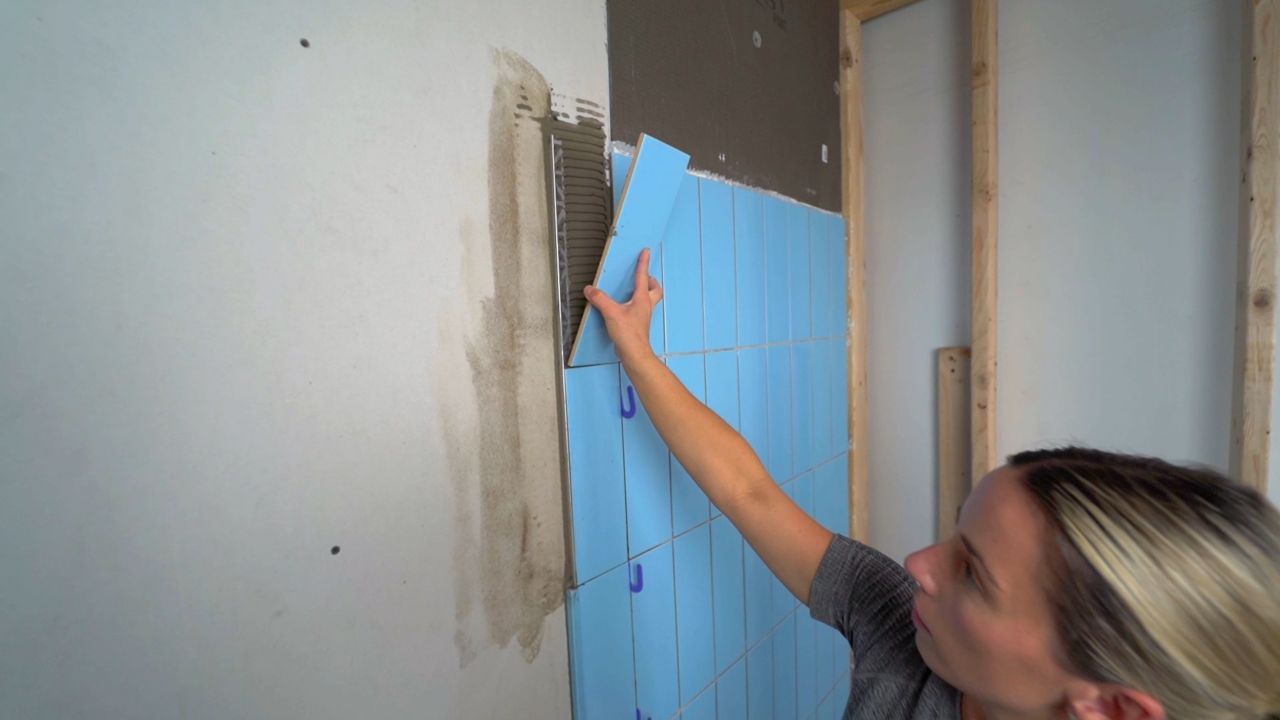 A person installing tile on a wall. A piece of metal profile trim is already embedded in the thinset, so the edge of the completed installation will have a clean, finished look.