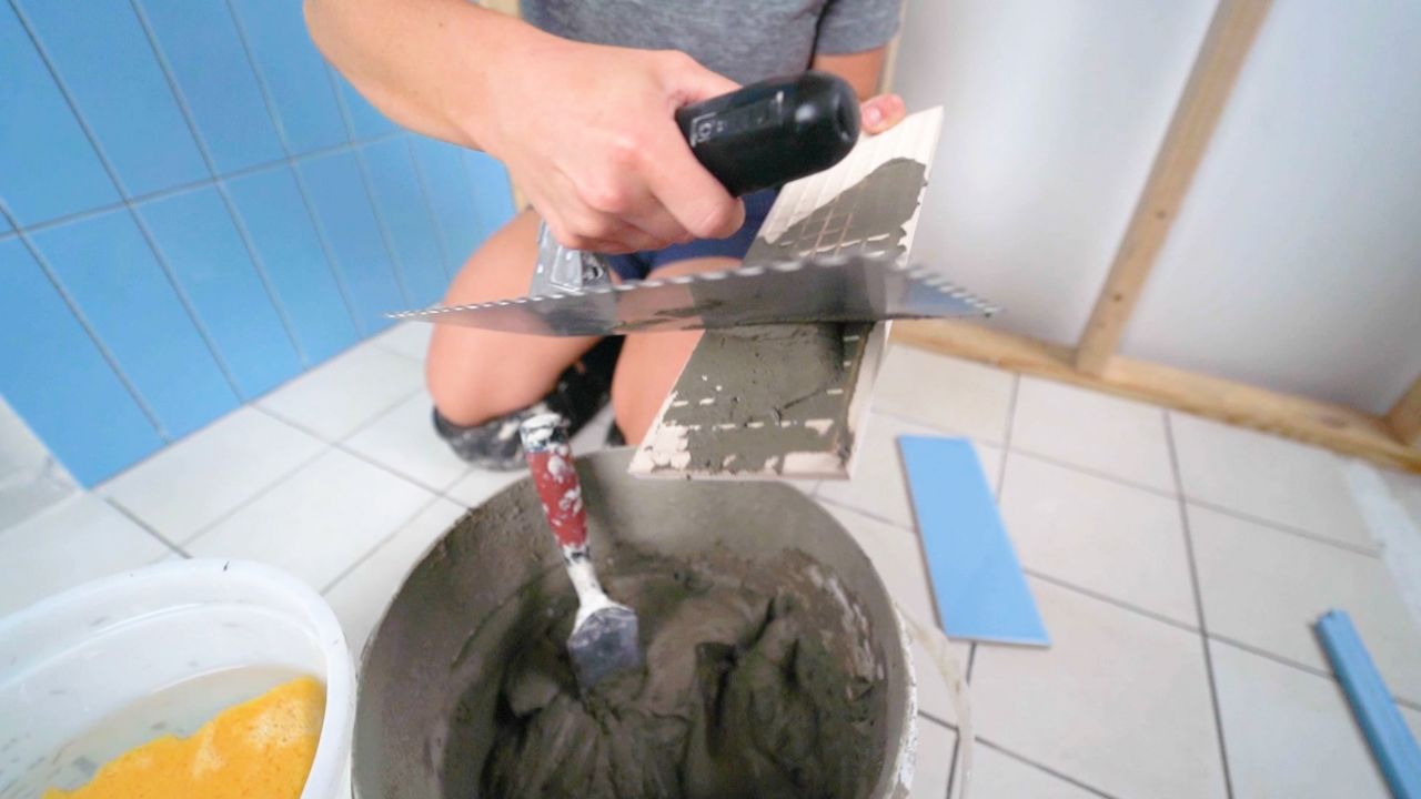 A person using a trowel to apply a skim coat of thinset mortar to the back of a tile prior to installation.