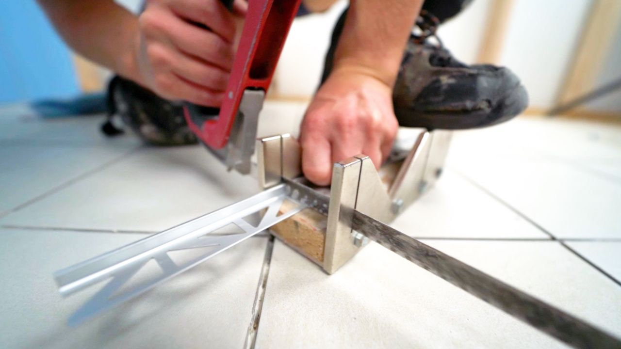 A person uses a miter box and saw to cut a piece of metal profile trim to the correct length.