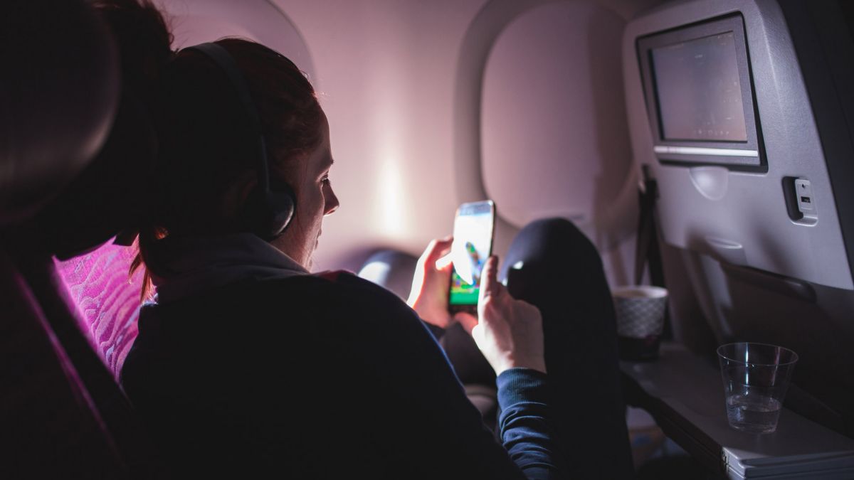 A person with headphones on curls up in an airplane seat and looks at something on a mobile phone screen. 