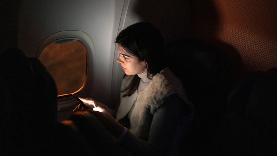 在昏暗的机舱里，一位留着长发的乘客坐在飞机窗户旁边, 眺望夕阳, 手机的光照亮了他们的脸.
