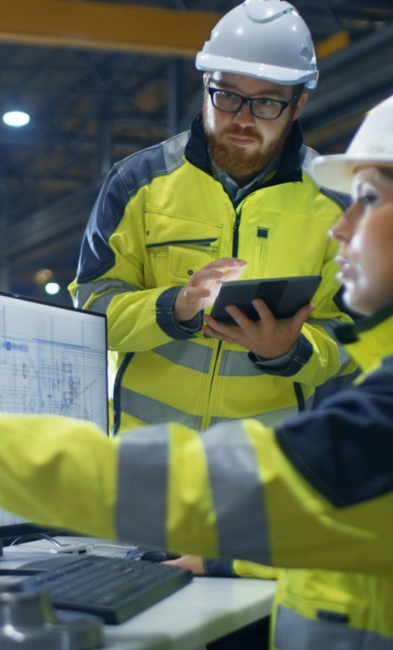 Workers in hard hats and high-visibility coats review information on computer and tablet screens.