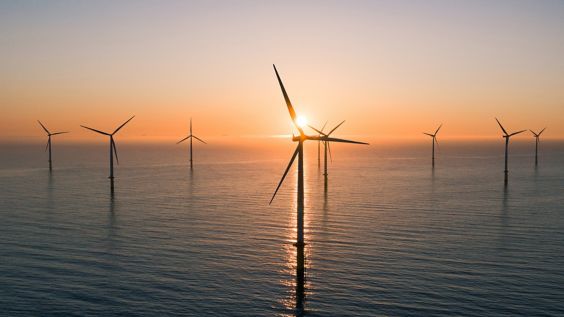 A group of wind turbines emerge from the ocean surface as the sun sets behind them.
