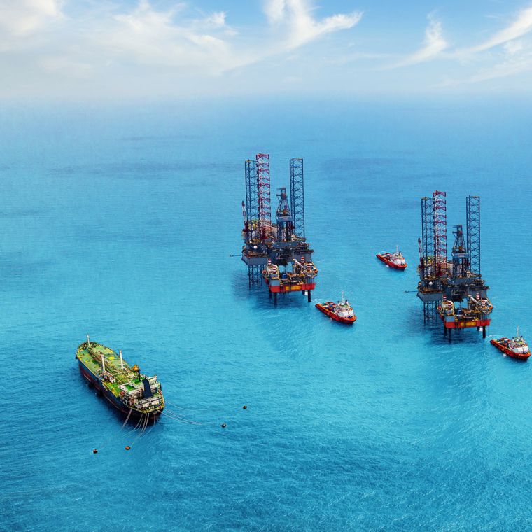 Two offshore oil rigs sit side by side, with boats nearby in a bright blue sea.
