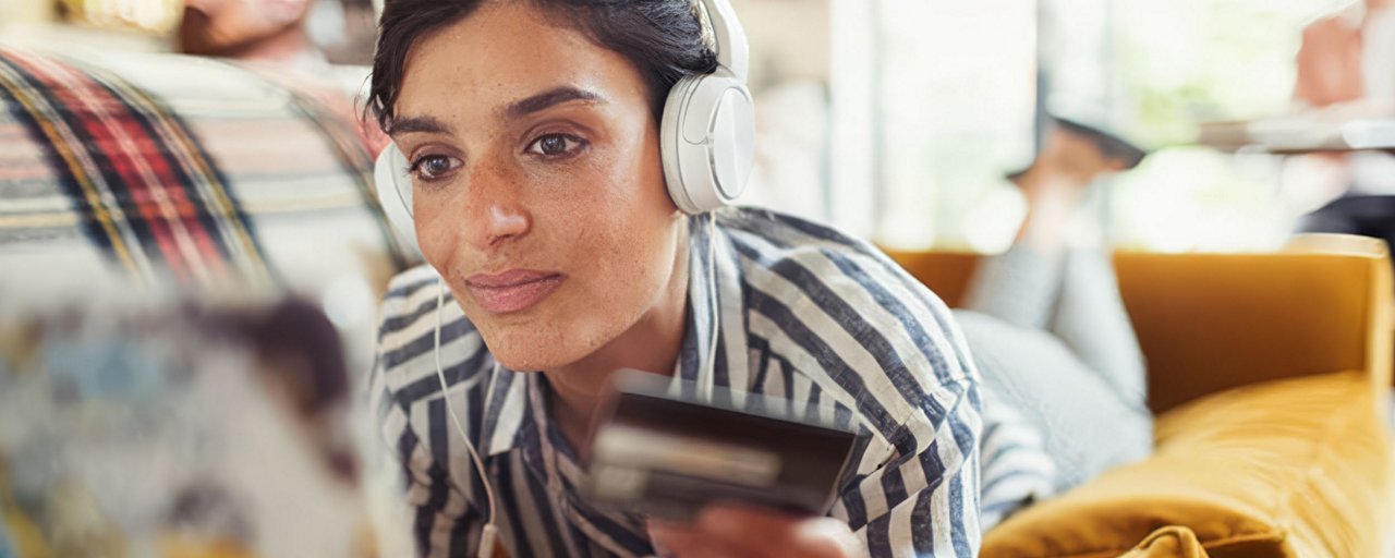 A young woman on her couch with headphones on, staring at her computer while holding a credit card in her hand