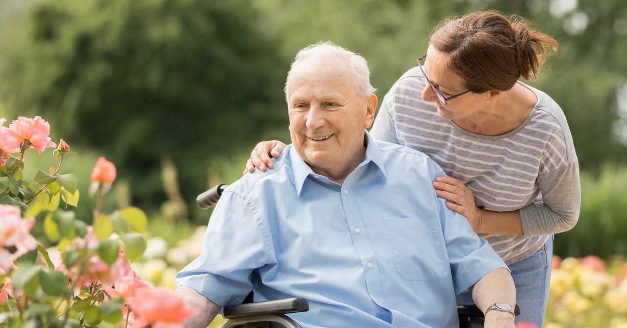 Older adult in a wheelchair outdoors with an adult family member providing support in a garden.