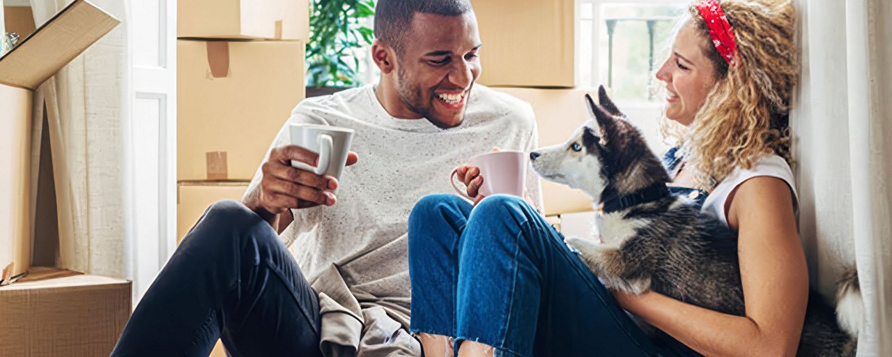 A couple drinking coffee with their husky puppy in a house full of moving boxes