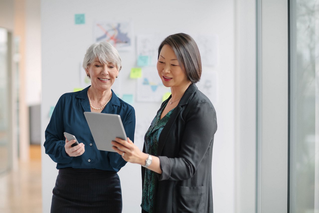 Two professional women stand together in a modern office, reviewing content on a digital tablet. One woman holds a smartphone while they discuss information displayed on the screen. The background shows bright windows and blurred wall notes, suggesting a collaborative workspace. The overall mood is professional, friendly, and focused on teamwork and technology.