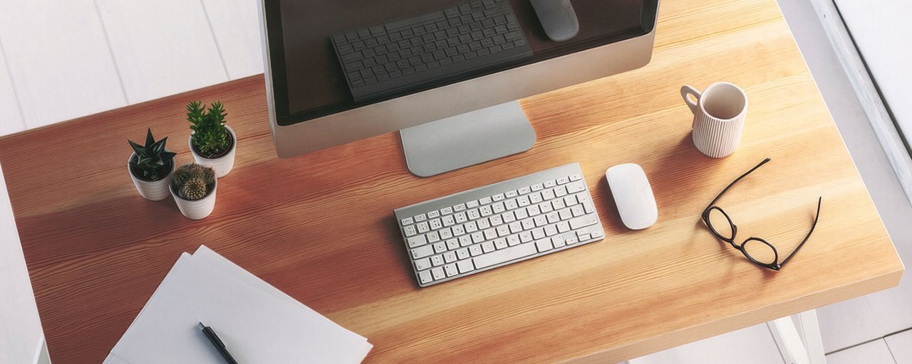 Overhead of a wooden desk with a monitor, keyboard, mouse, papers, coffee, and a pair of glasses on it