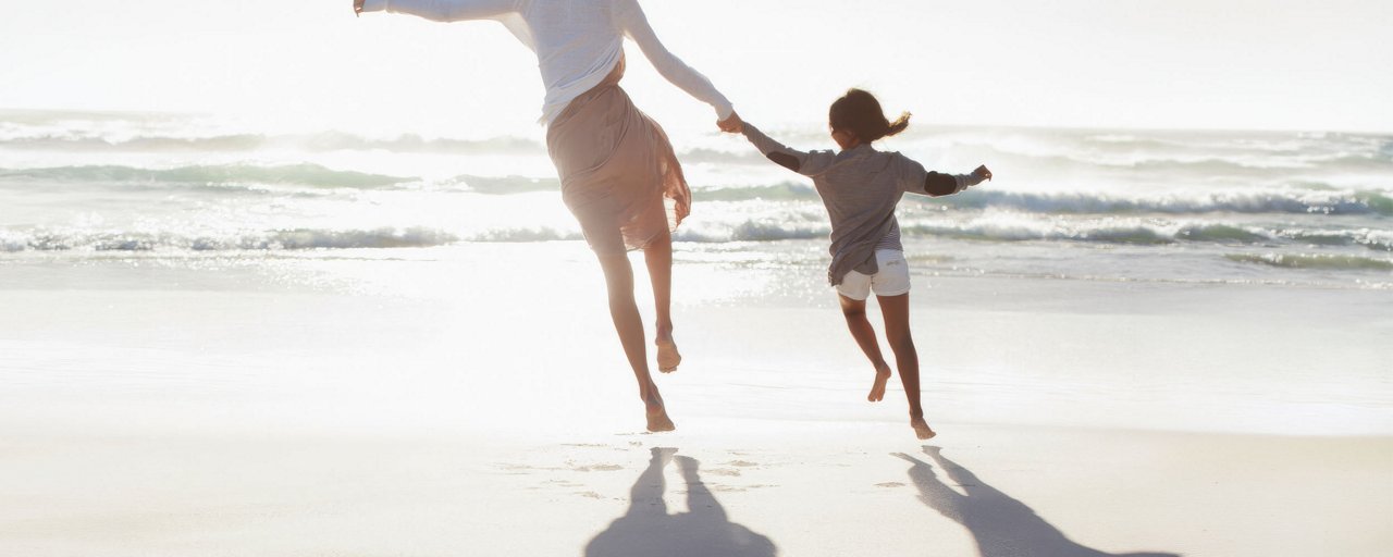 A mother and her young daughter holding hands and running on the beach at sunset