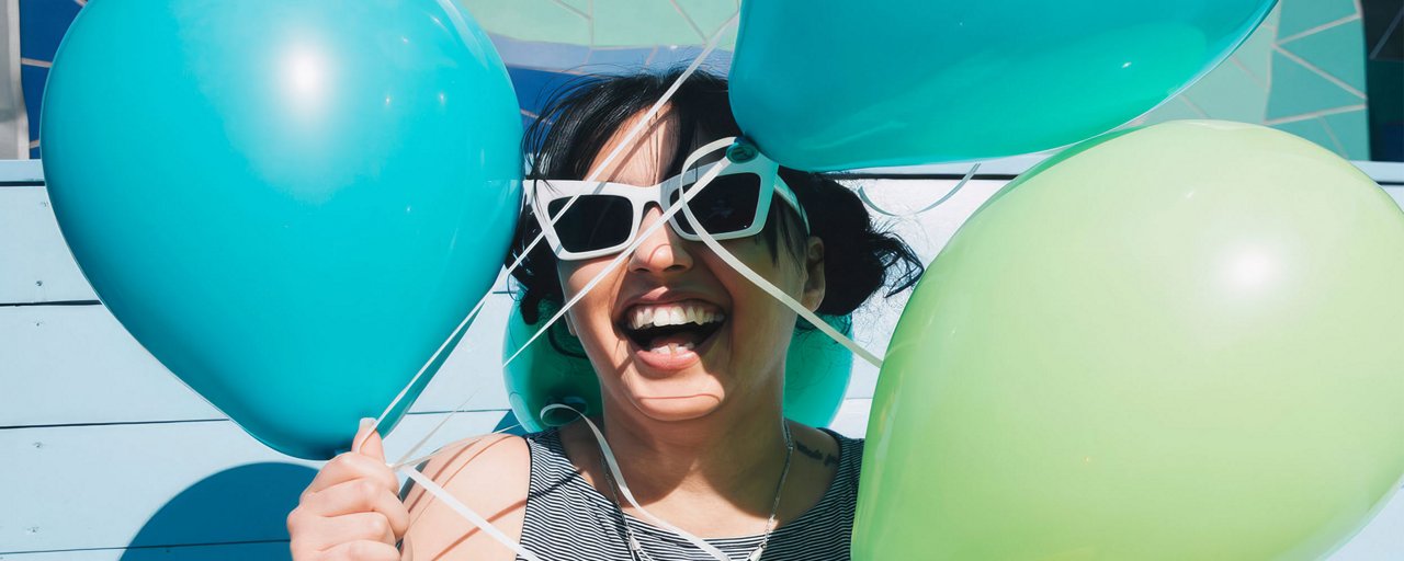 A woman in white sunglasses laughing against a blue house holding a bundle of balloons 