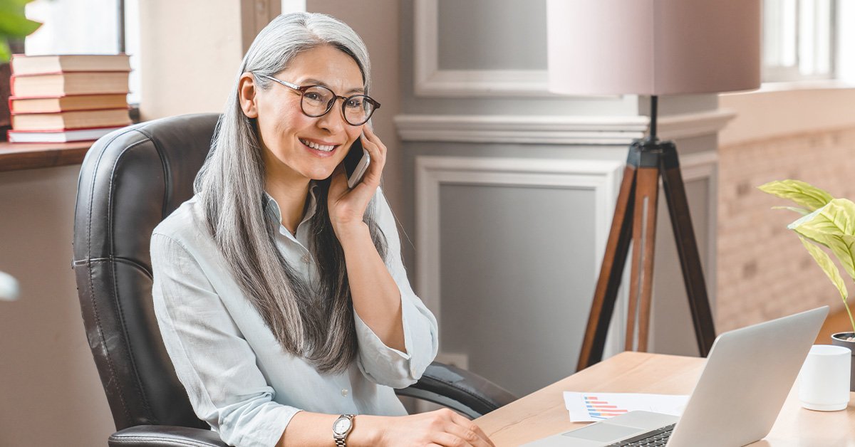 Femme  mûre souriante et assise avec un ordinateur portable sur le bureau devant elle à la maison 
