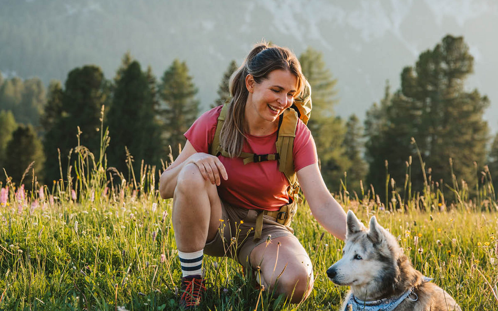 Person kneeling in a grassy meadow, petting a dog with mountains and trees in the background.