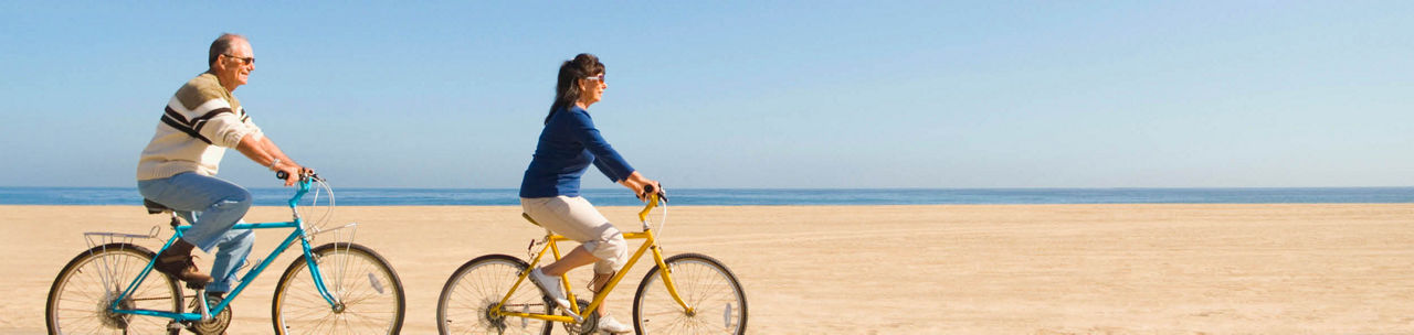 Two people riding bicycles side by side on a sandy beach with the ocean and clear blue sky in the background.