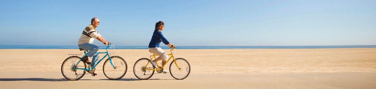 Two people riding bicycles side by side on a sandy beach with the ocean and clear blue sky in the background.