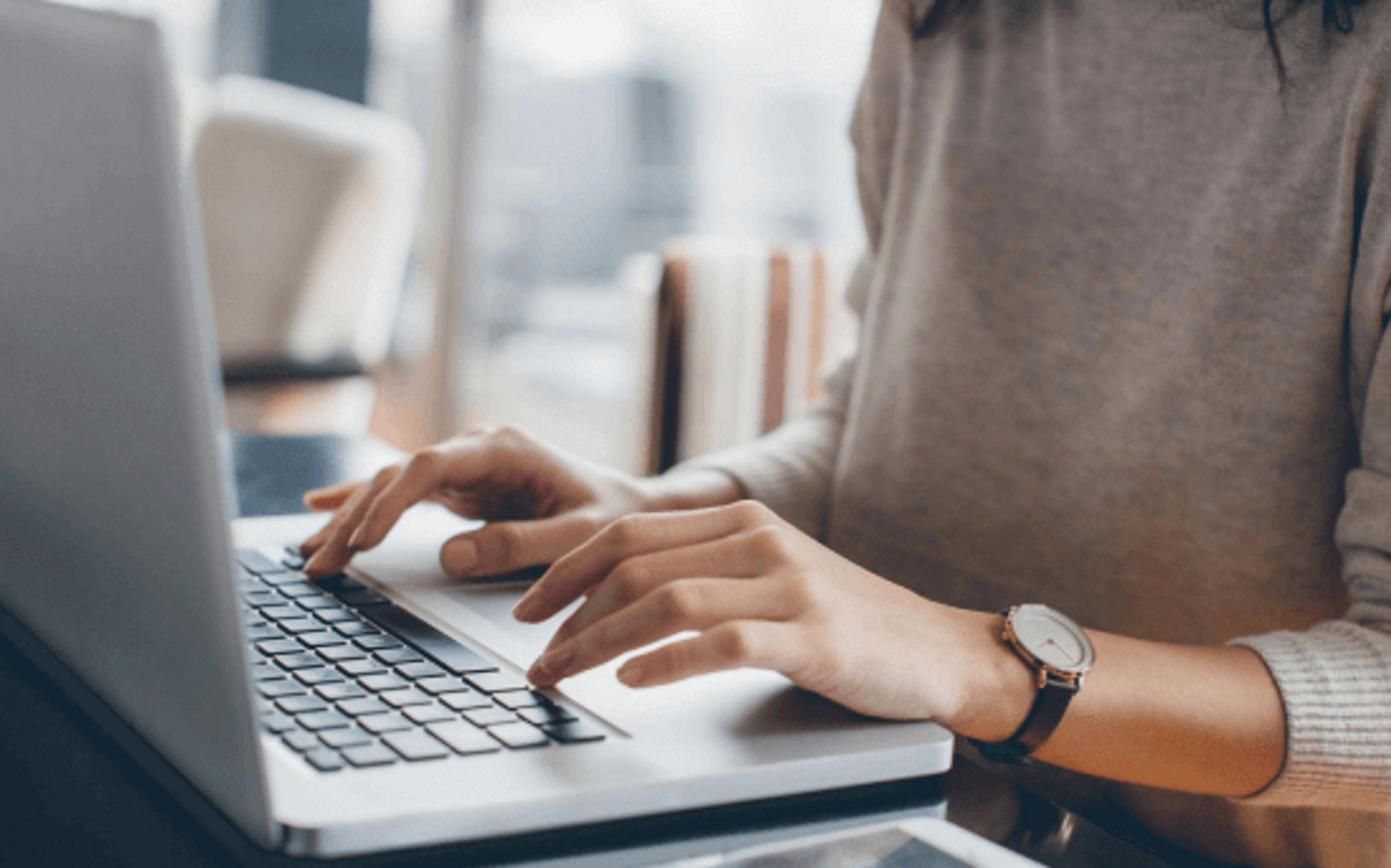 Person typing on a laptop keyboard at a desk, wearing a gray sweater and wristwatch in a modern workspace.