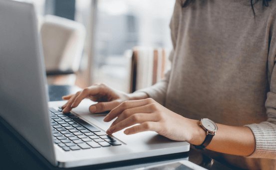 Person typing on a laptop keyboard at a desk, wearing a gray sweater and wristwatch in a modern workspace.