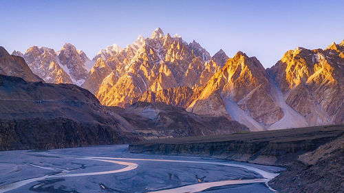 A serene mountain landscape with a snow-capped peak reflected in a tranquil lake under a clear blue sky and soft clouds.