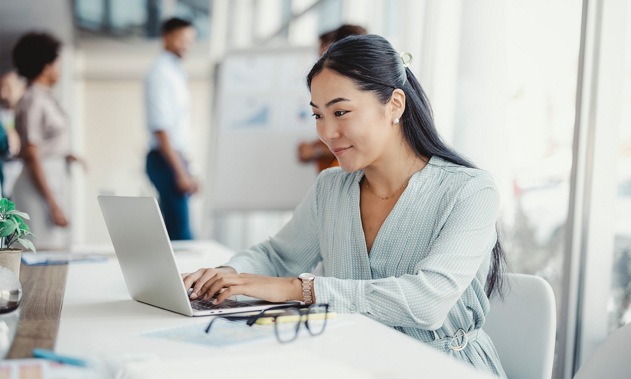 A businesswoman sits at her desk, smiling as she works on her laptop in a professional office environment.