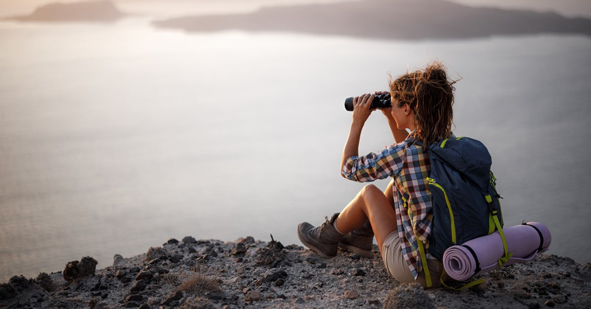 Person sitting on rocky shore looking through a pair of binoculars at an ocean vista dotted with craggy land masses and a brightening sky on the horizon