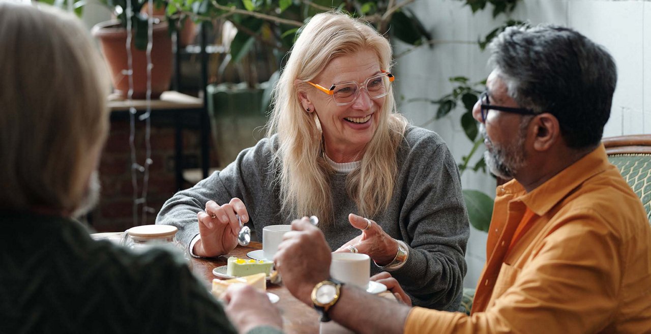 Trois personnes âgées assises à une table dans une pièce lumineuse, partageant un café et un dessert tout en discutant, avec des plantes d’intérieur et de la lumière naturelle en arrière-plan.