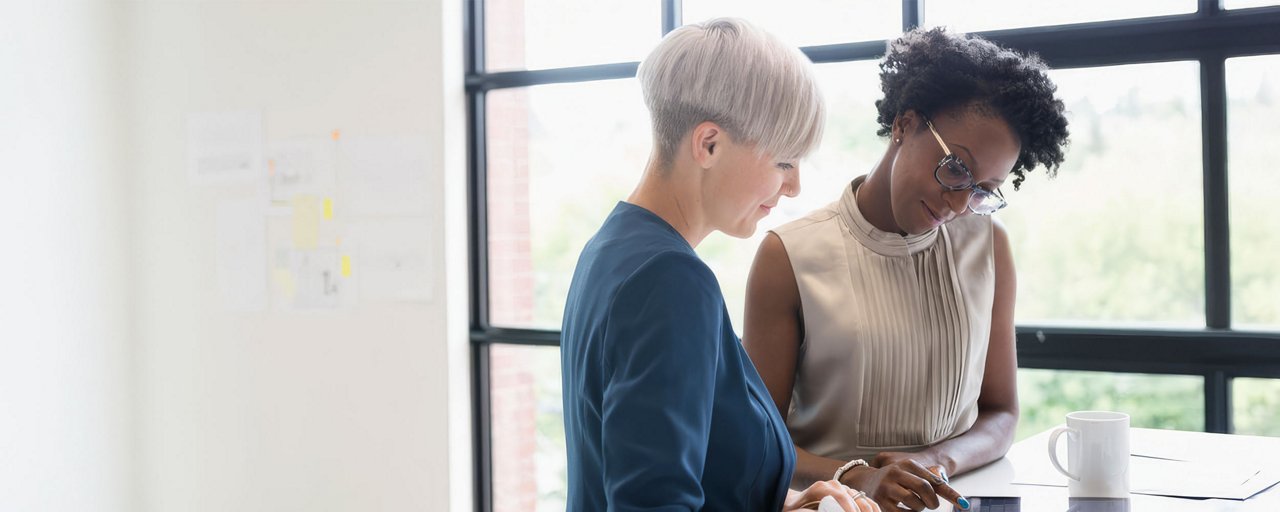 Two women working together on a tablet in an office space at a white table