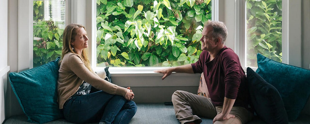 A man and woman sitting in their house on a window seat in front of a wall of ivy