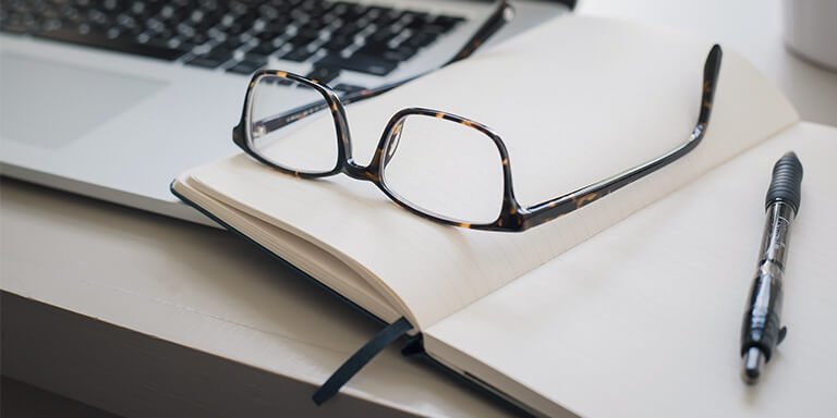 Close up of a pair of glasses resting on an open notebook next to a laptop computer and a pen 