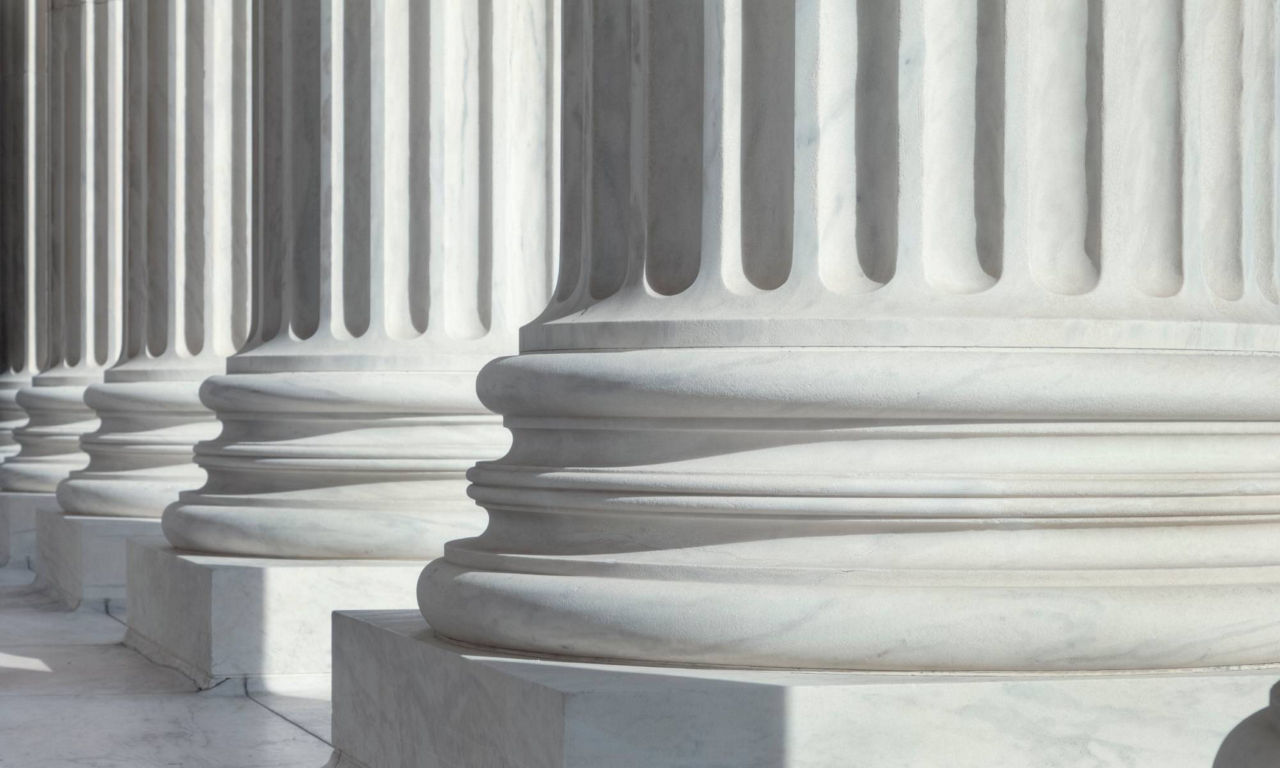 Close-up view of large marble columns showcases their fluted shafts and sturdy bases. The architectural elements are bathed in soft daylight, creating strong shadows and highlights across the stone surface. The scene suggests a formal institutional or governmental building with neoclassical design. Smooth textures and pale tones emphasize symmetry, stability, and permanence.