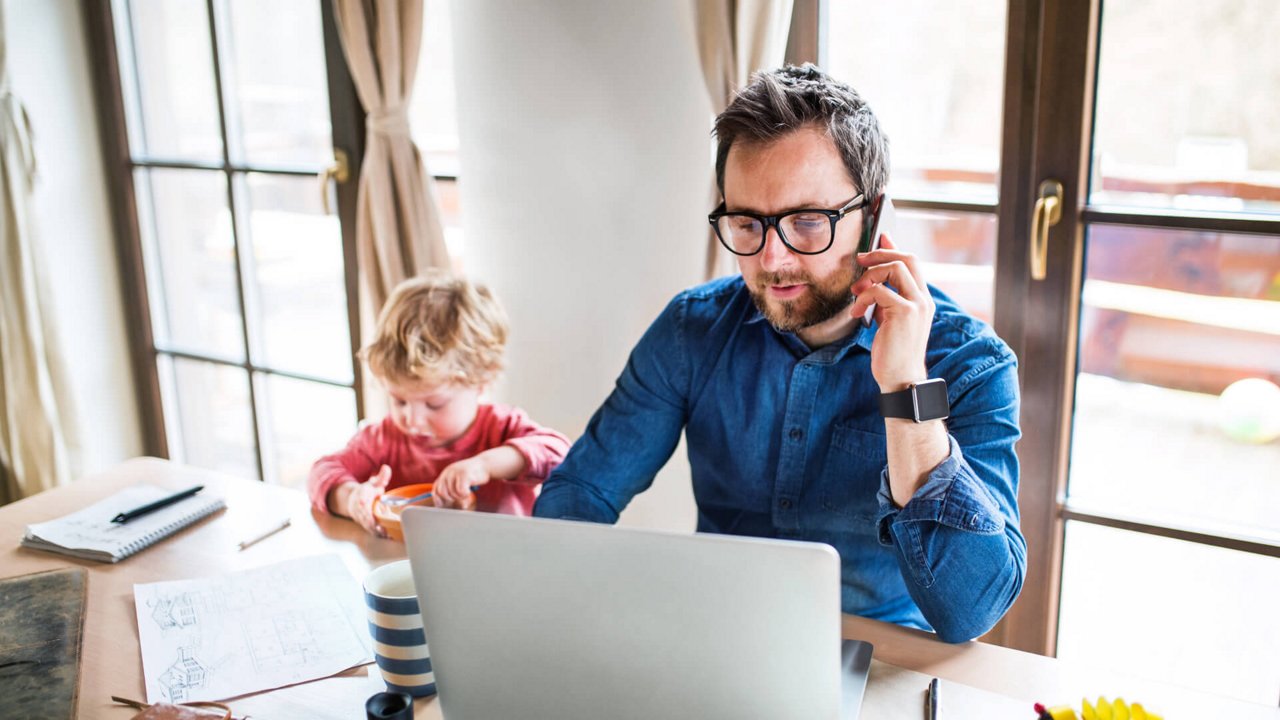 Person working on a laptop and phone at a home table while a young child sits nearby.