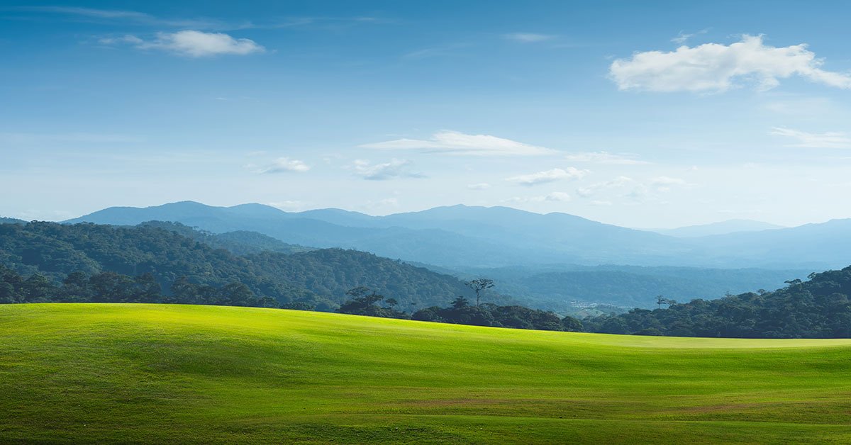 Panoramic landscape of wide open rolling green fields, hills/mountains, and blue sky with a few clouds