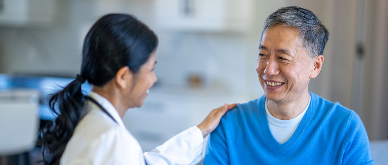 A senior gentleman of Asian decent, sits in the comfort of his own home with his doctor, during a Home Care visit.  His doctor is dressed professionally and is taking notes electronically.