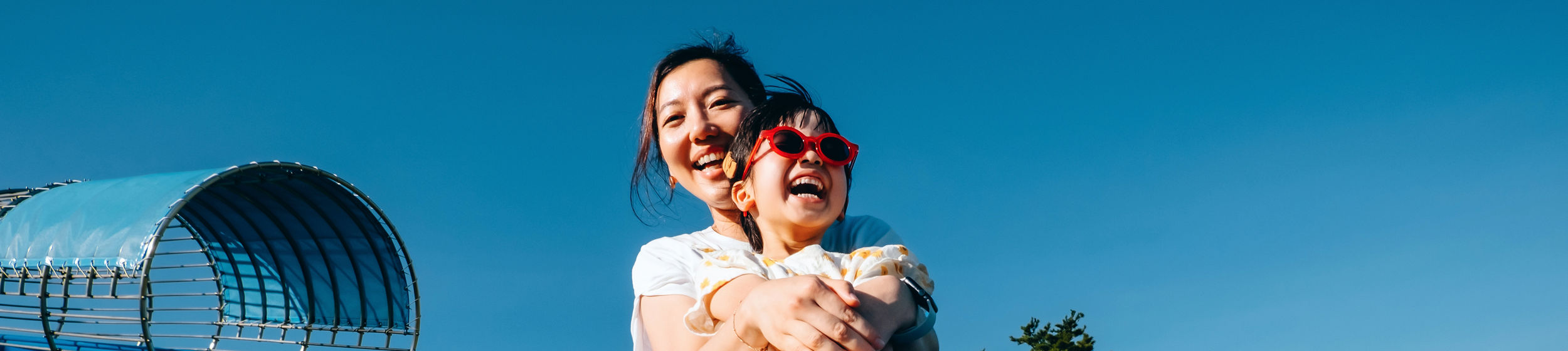 Happy young Asian mother with her little daughter having fun playing on the tube slide together in an outdoor playground on a lovely sunny day. Outdoor fun. Happy childhood and motherhood. Mother and daughter bonding moment. Happy family life