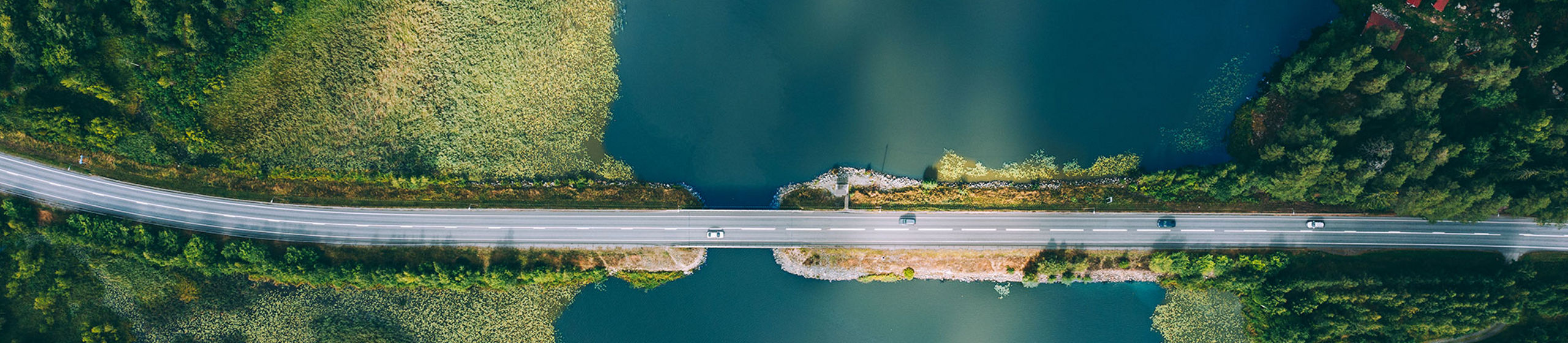 An aerial view shows a straight road bridge crossing a calm blue-green lake. Dense evergreen forest surrounds both sides of the water, creating a strong contrast between land and lake. The water reflects the sky, adding soft cloud patterns to the scene. The composition emphasizes symmetry and the isolation of the roadway cutting through the natural landscape.
