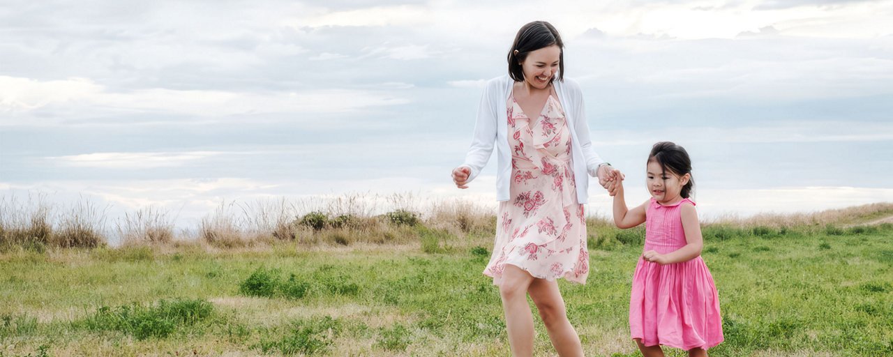 A woman and her young daughter in pink dresses running together in thick grass