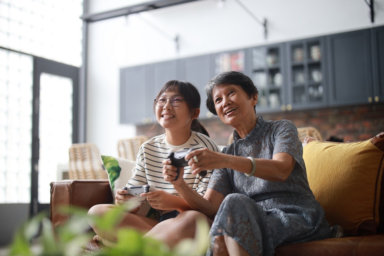 A cheerful Asian grandmother and his granddaughter are sitting on the sofa in the living room, having fun playing video games together at home. This heartwarming scene embodies the happy retirement concept