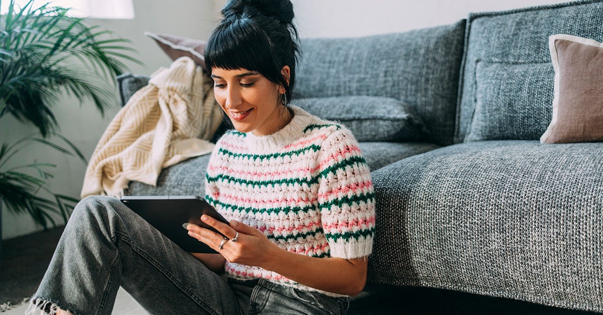 A young woman is comfortably seated on a sofa at home, engrossed in a book while her laptop sits nearby.