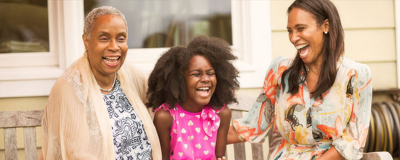 A grandmother, mother, and daughter laughing together on a bench outside of their house