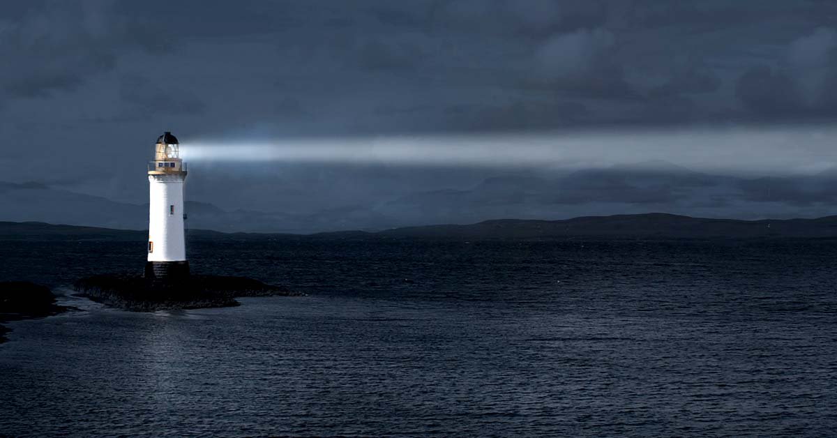 A serene sunset paints the sky blue as the lighthouse beacon illuminates the coastal beach and the blue sea.