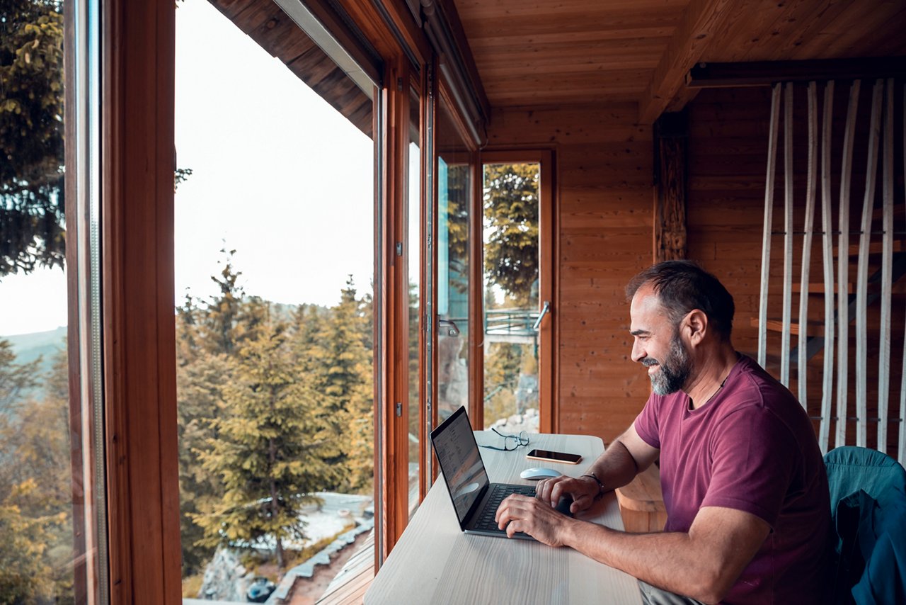 A man works on a laptop inside a wooden cabin with large windows