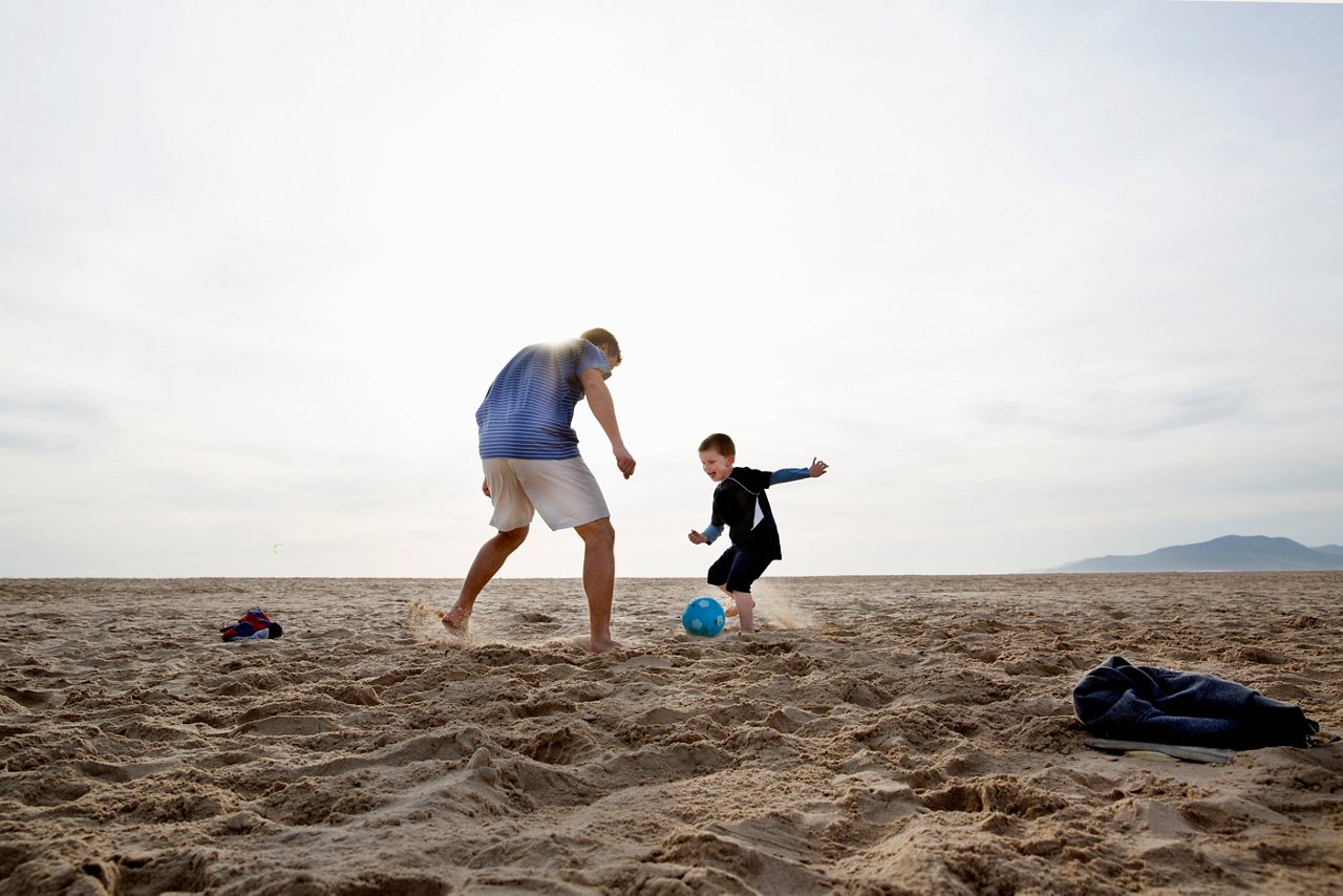 Two people play soccer together on a sandy beach with a blue ball
