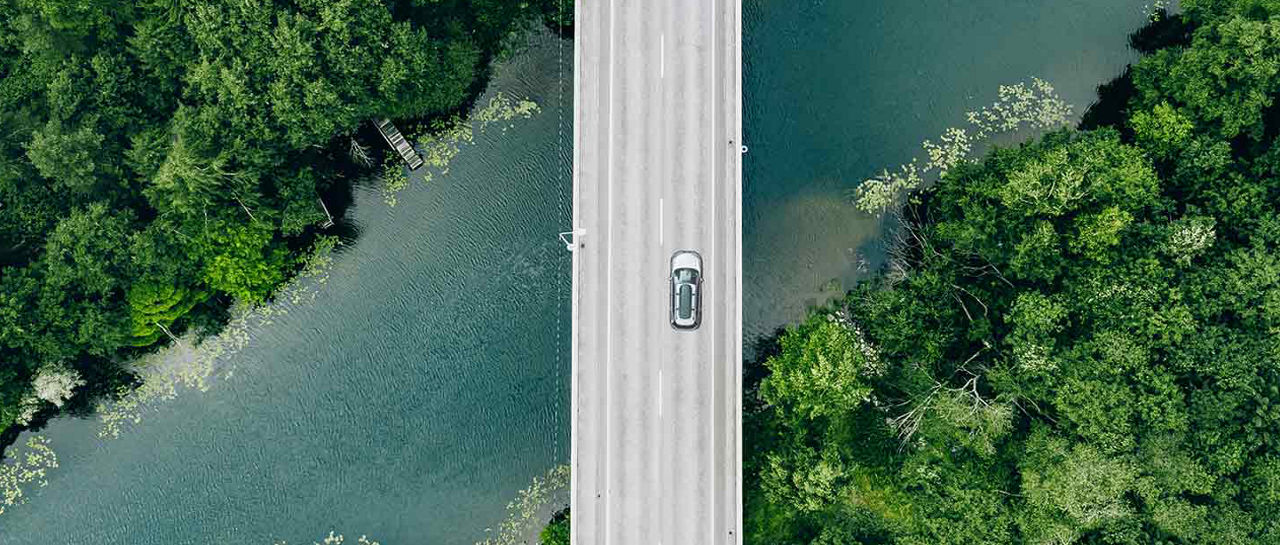 An aerial view shows a single car driving across a long, straight bridge. The bridge spans a calm river that winds through dense green forest on both sides. Lush trees and foliage create a vivid contrast with the muted color of the water and the light-toned roadway. The scene emphasizes solitude, nature, and the intersection of infrastructure with a natural landscape.