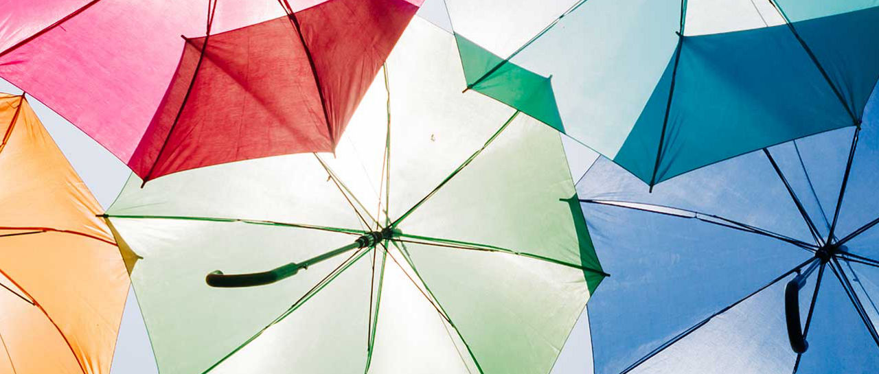 A collection of open umbrellas in various bright colors fills the frame. The umbrellas are viewed from below, creating a canopy effect against a clear blue sky. Strong sunlight highlights the vivid hues and casts subtle shadows on the fabric. The image conveys a playful, cheerful atmosphere with a focus on color and pattern.