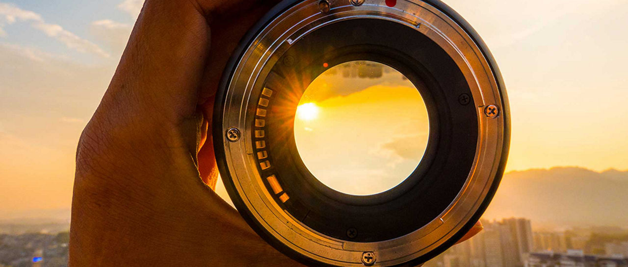 A hand holds a camera lens up to the sky, framing the setting sun through the glass. The city skyline and distant mountains appear blurred in the background, emphasizing the circular lens opening. Warm golden light from the sunset reflects off the metal edges of the lens. The image highlights themes of photography, perspective, and urban exploration.