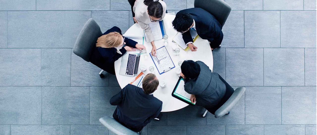 A group of five business professionals sit around a round white table in a modern office setting. They are engaged in a meeting, reviewing documents, notes, and digital devices. Laptops and a tablet are visible on the table, suggesting collaboration and data sharing. The scene is viewed from above, with cool gray floor tiles creating a clean, corporate atmosphere.