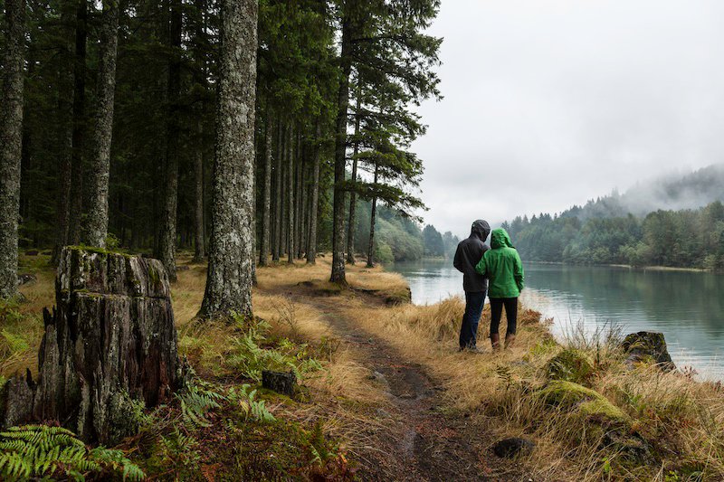 A couple wearing raincoats looks out over a foggy, misty lake in the fall in an evergreen forest in Washington state.