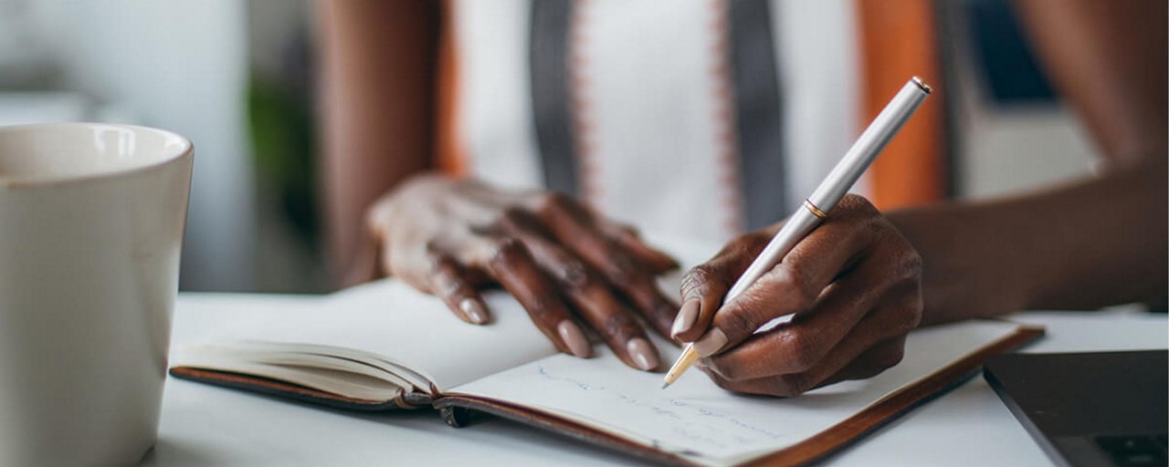 Close up of a woman writing in her open journal with a cup of coffee sitting on the table 